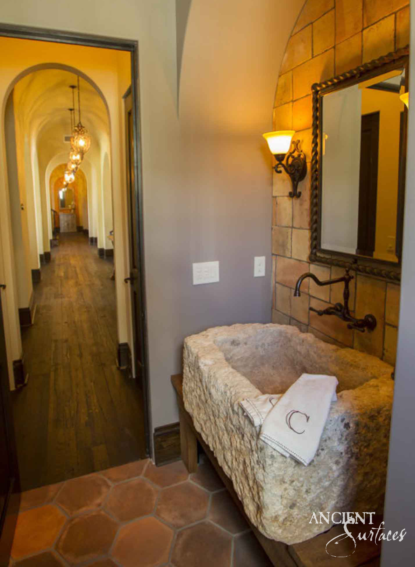 Deep reclaimed antique limestone trough sink from Ancient Surfaces with thick rough quarried walls and an uneven rim, raised on a timber cradle below a wall mounted bronze tap in a terracotta tiled powder room