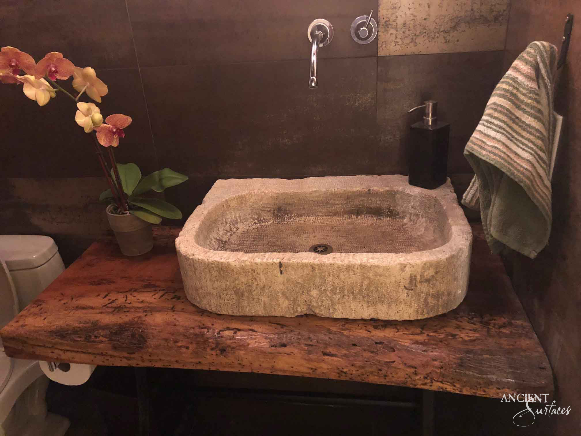Reclaimed antique limestone vessel basin from Ancient Surfaces with rounded corners and a shallow point tooled bowl, sitting on a live edge timber counter below a wall mounted chrome spout