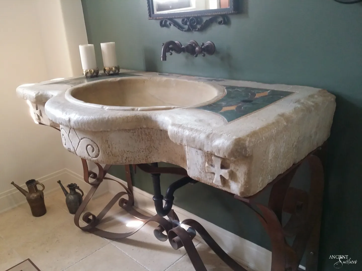Reclaimed antique limestone slab sink from Ancient Surfaces with a shaped front edge, a wide oval basin, a green stone inlay panel set into the deck and a carved cross on the apron, standing on a scrolled iron base