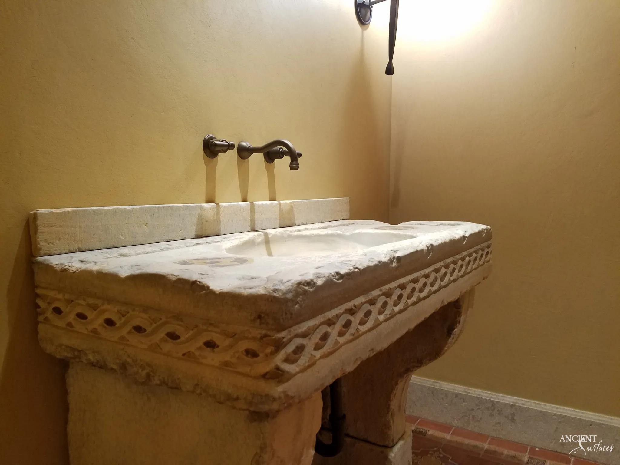 Reclaimed antique limestone console sink from Ancient Surfaces with an interlaced ribbon band carved along its apron, a shallow rectangular basin sunk into the deck and a separate limestone backsplash slab behind