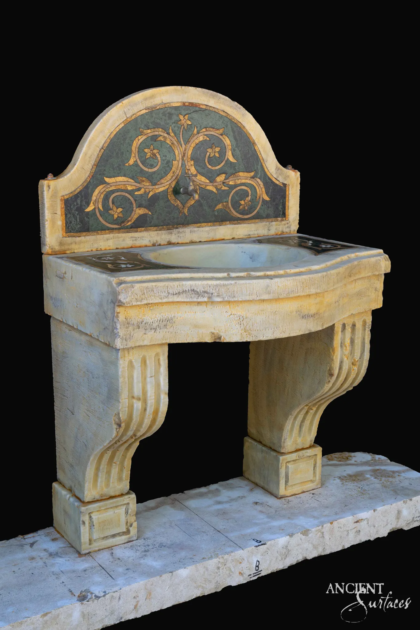 Reclaimed antique limestone sink console from Ancient Surfaces photographed against black, with an arched backsplash inlaid in green and ochre foliate scrollwork, an oval basin and two fluted scroll corbel legs
