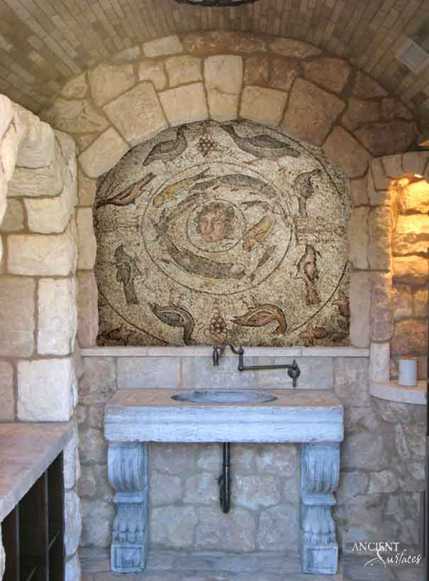 Reclaimed antique limestone console sink from Ancient Surfaces, a thick grey slab with a shallow round basin dished into the deck, carried on two carved acanthus corbels in a vaulted stone alcove