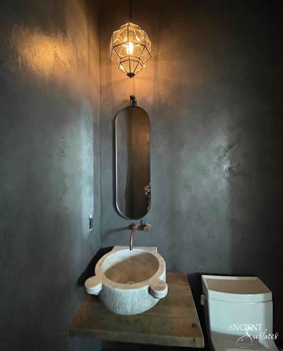 Round reclaimed antique limestone vessel bowl from Ancient Surfaces with thick faceted walls and two carved lug handles, set on a reclaimed timber shelf below a wall mounted spout