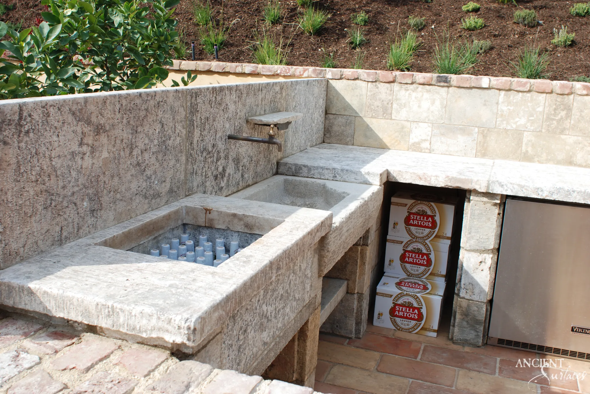 Outdoor reclaimed antique limestone sink station from Ancient Surfaces with a deep rectangular basin beside a smaller shallow one, a tall limestone backsplash slab and a bronze bibcock on a carved stone bracket