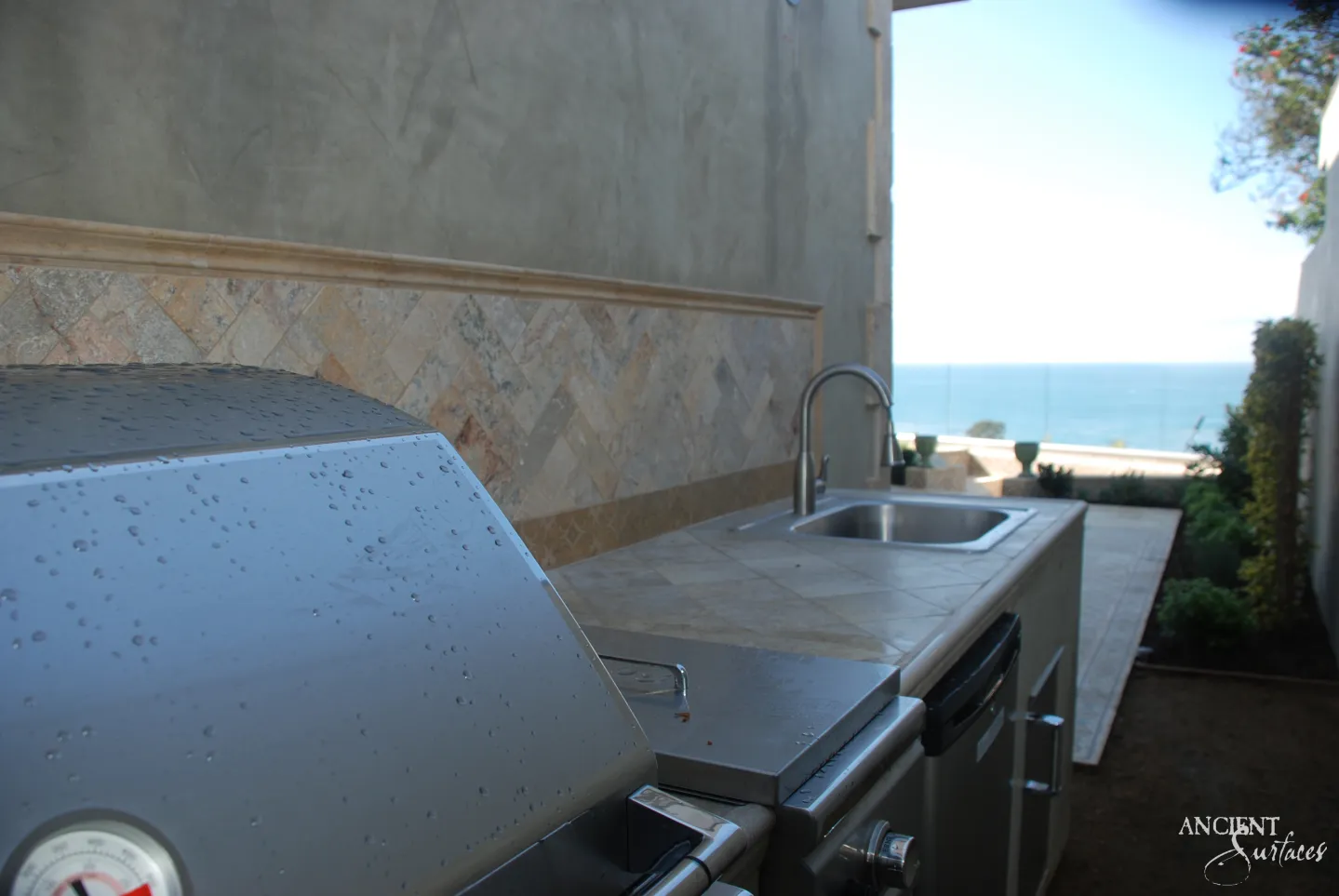 Reclaimed antique limestone counter from Ancient Surfaces laid as honed pale grey tiles around an outdoor kitchen sink, with a warm gold split face limestone backsplash band capped by a moulded rail
