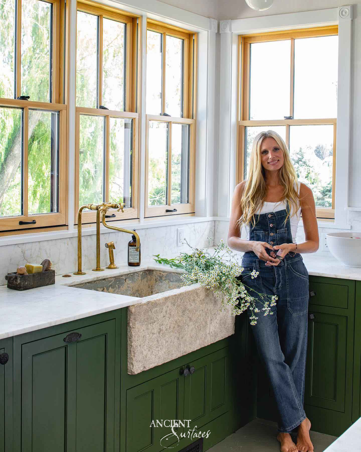 Reclaimed antique limestone farmhouse sink from Ancient Surfaces set into deep green cabinetry, a single deep basin with a coarse cream apron front standing proud of the marble counter beside a brass bridge tap