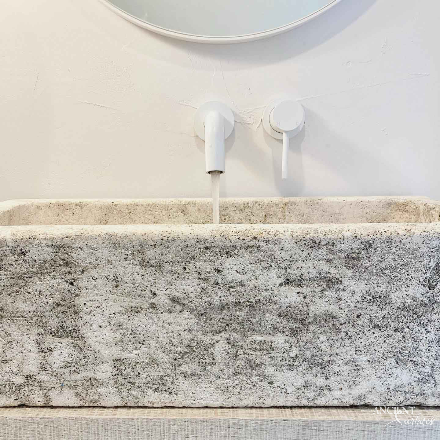 Close view of a reclaimed antique limestone trough basin from Ancient Surfaces, its deeply pitted grey and cream front face below a flat even rim, set on a pale timber shelf under a white wall spout